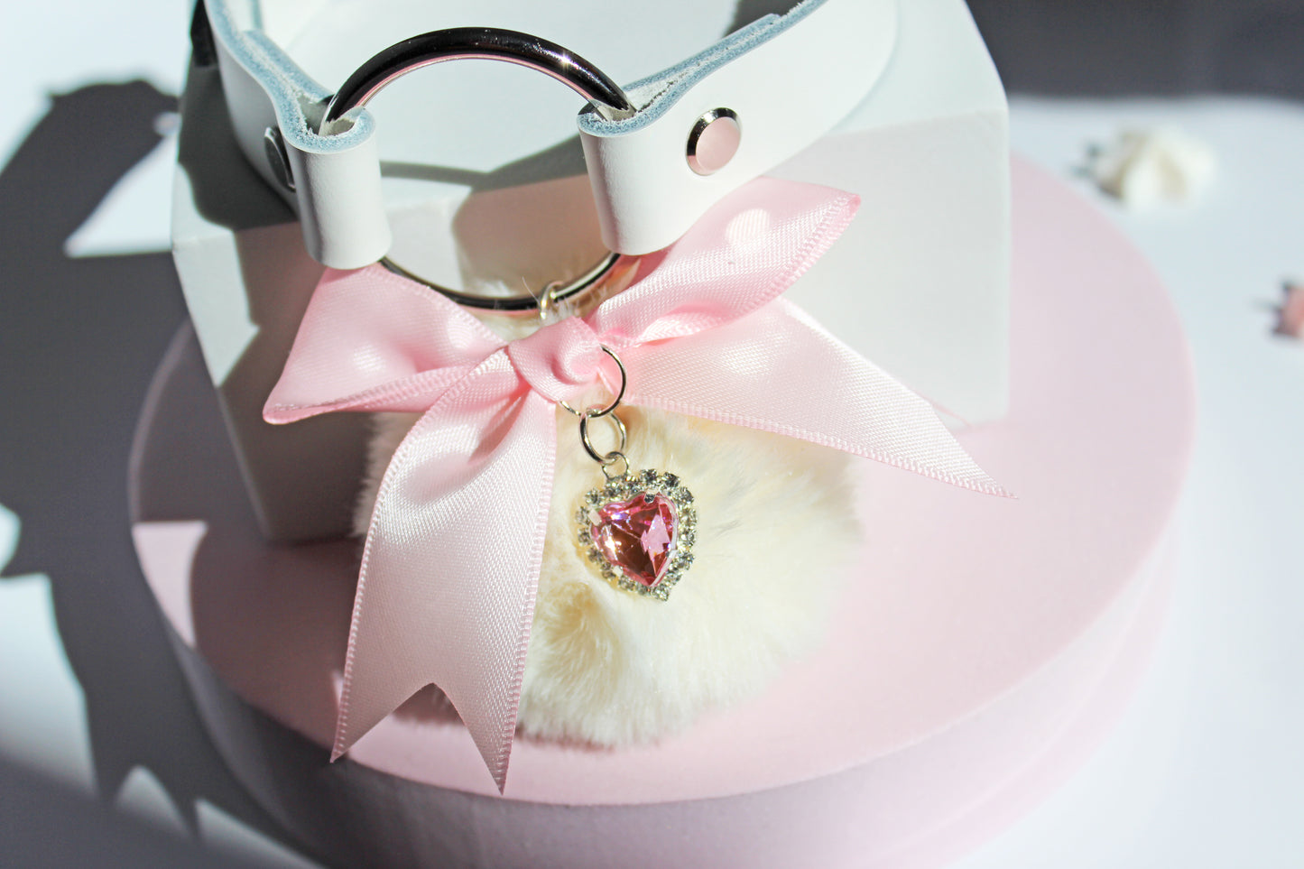 Kawaii white pom collar with a pink bow, heart, and white pom on a pink stand with a white background.