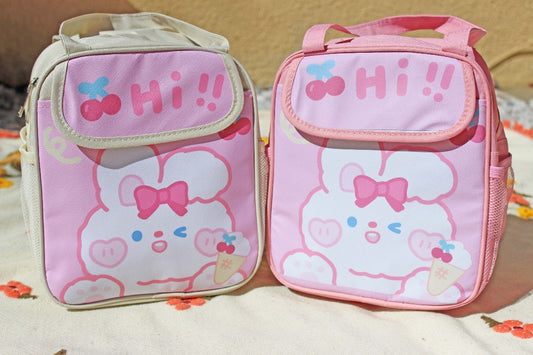One white and pink lunch bag with a cute bunny eating ice cream next to an all pink lunch bag with a bunny eating ice cream on set on a blanket outside.