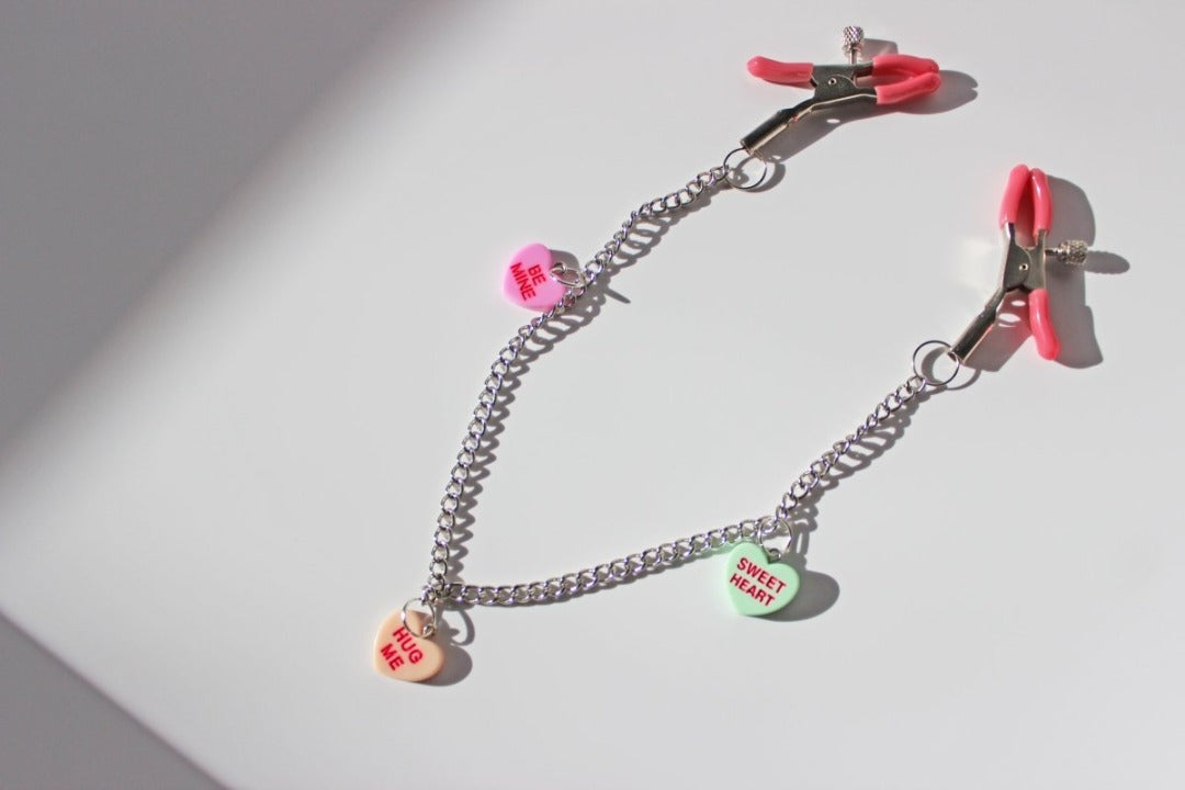 Pink PVC capped silver tension screw nipple clamps connected by a chain with three candy heart charms on them placed on a white background.