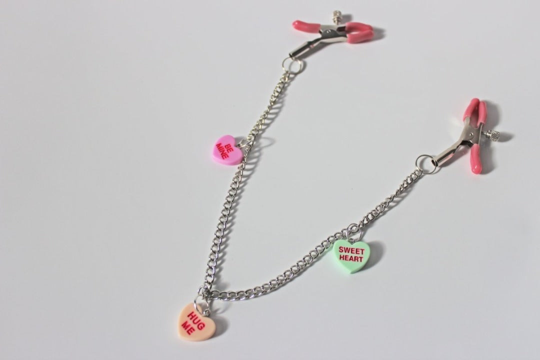 Pink PVC Capped silver tension screw nipple clamps connected by a chain with three candy heart charms on them placed on a white background.