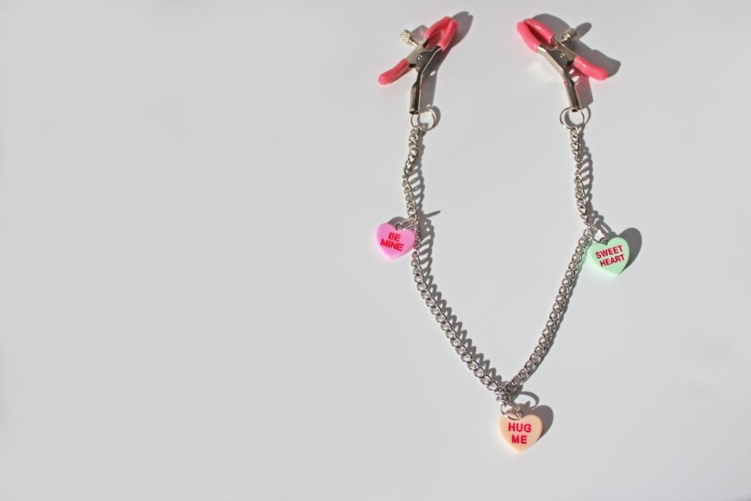 Pink PVC Capped silver tension screw nipple clamps connected by a chain with three candy heart charms on them placed on a white background.