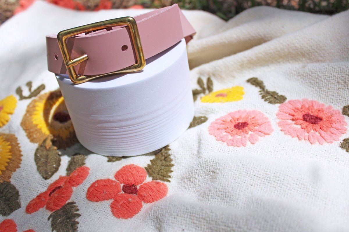 Pink leather rose submissive collar turned backwards to show the gold buckle hardware on a white stand with a blanket.