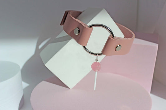 Pink leather lollipop submissive collar on a white  block with pink shapes and a white background.