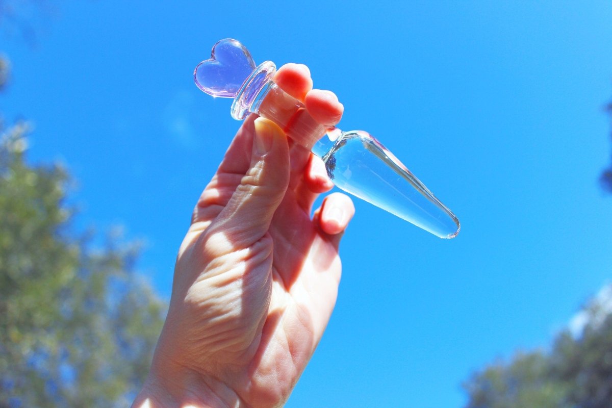 tapered clear butt plug with a pink heart base being held up in front of the sky